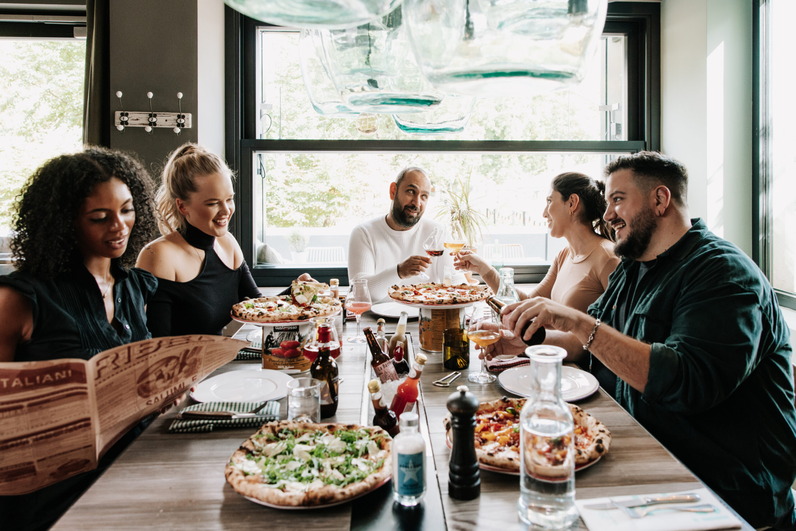 A group of 5 people is enjoying lunch on a large table, there are several different kinds of pizza as well as dirnks on the table