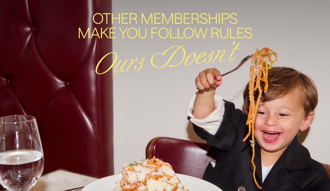 A child in a black jacket playfully holds spaghetti on a fork, with an elegant dining setting in the background.