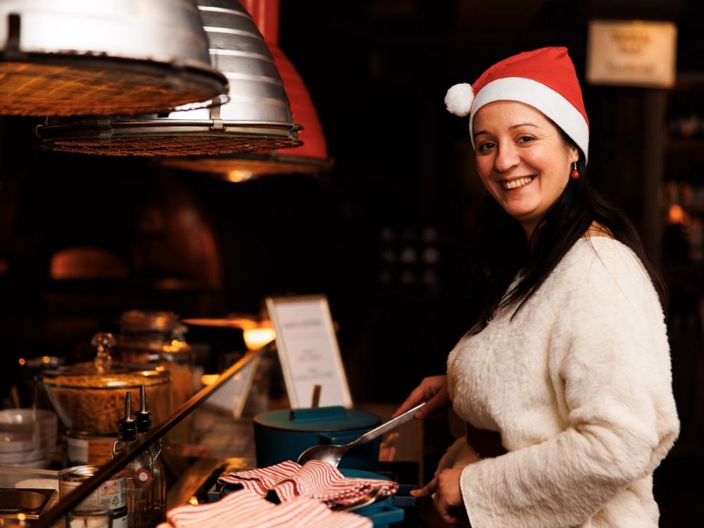 Eine Frau mit Weihnachtsmütze und kuscheligem weißen Pullover lächelt, während sie in einer warm beleuchteten Küche kocht. Festliche, fröhliche Atmosphäre mit Weihnachtsdekoration.