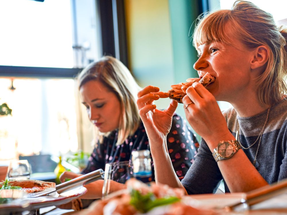 Eine blonde Frau beißt gut gelaunt in ein frisches Stück Pizza mit Parmaschinken, während sie mit einer Freundin an einem hellen Restauranttisch speist.