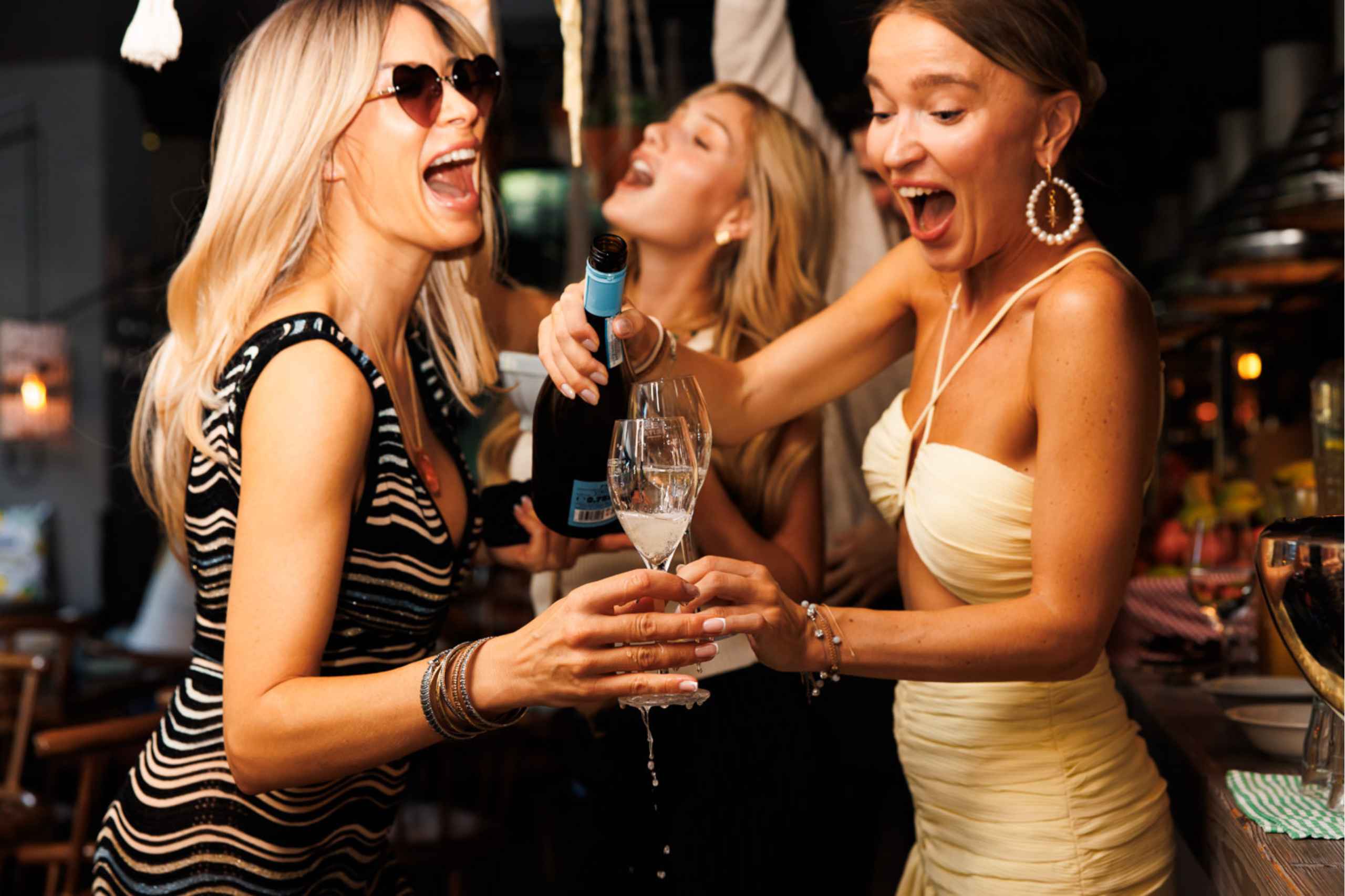 Drei ausgelassen feiernde Frauen lachen und stoßen mit Champagner an, während eine Flasche Schaumwein direkt in die Gläser eingeschenkt wird.