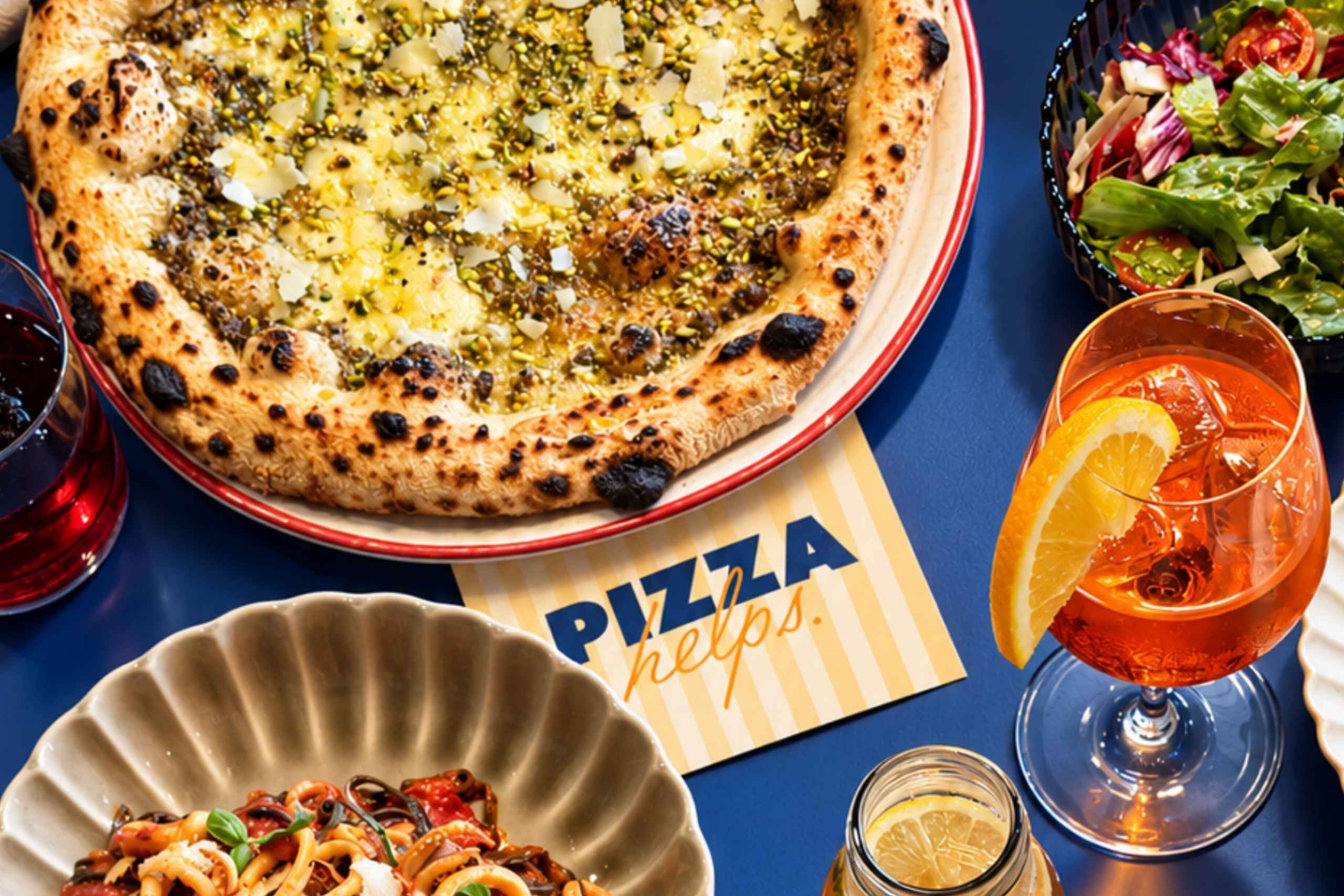 Ein farbenfroher Tisch mit einer großen Pesto Pizza mit Pistazien und Parmesan, Meeresfrüchte-Pasta, einem Aperol Spritz und einem Beilagensalat auf einem blauen Tisch.