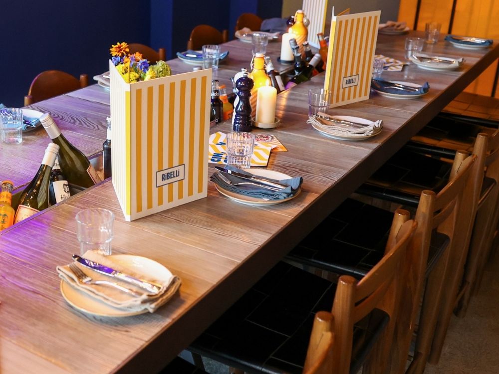 Long wooden dining table set for group dining at Ribelli Italian restaurant, featuring yellow striped menus, built-in bottle coolers, and candles.