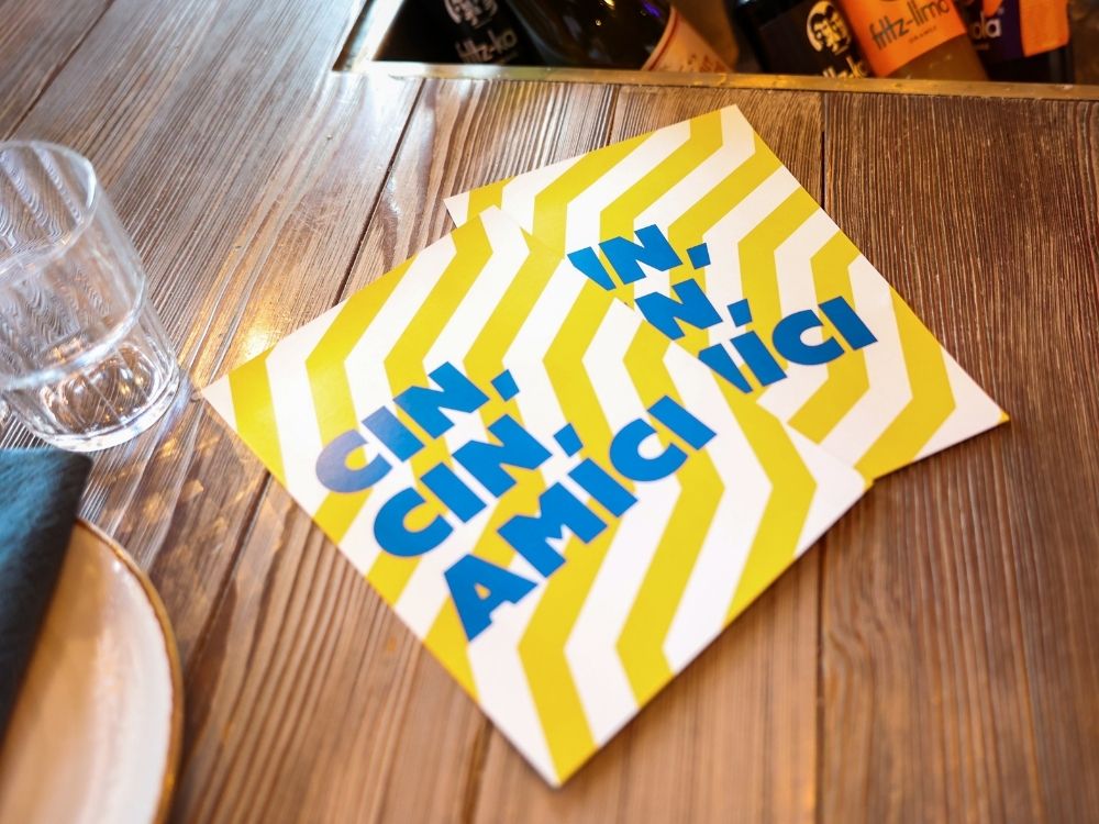 Custom yellow and white striped card with Italian text Cin Cin Amici Miei on a wooden table at Ribelli restaurant.